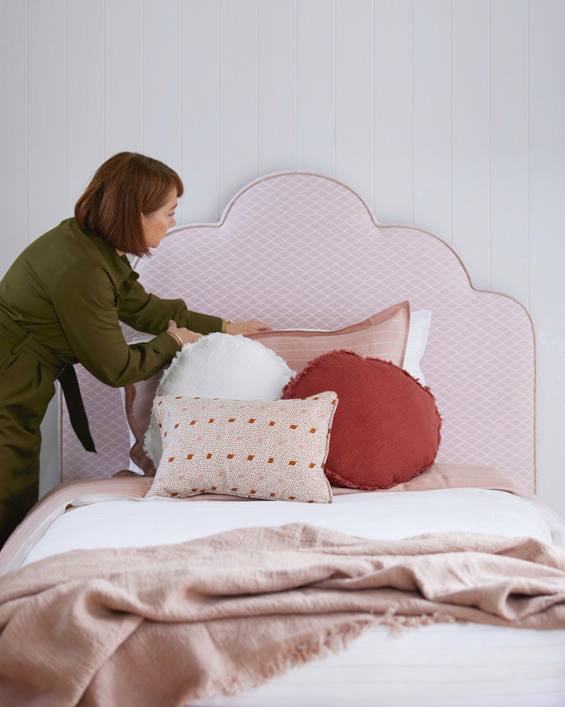 A styled kid's bedroom with a pink scalloped shaped bedhead. The bed shows white bedding and a lady arranging the pillowcases and cushions.