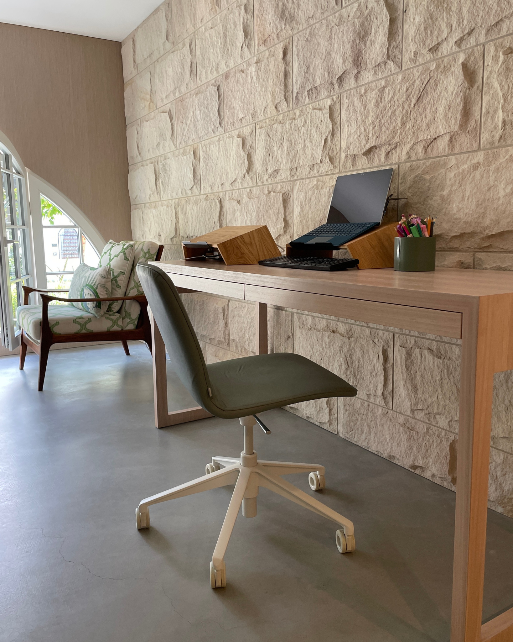 A modern home office setup featuring a light wood study desk paired with a dark green swivel office chair, set against a textured stone wall.