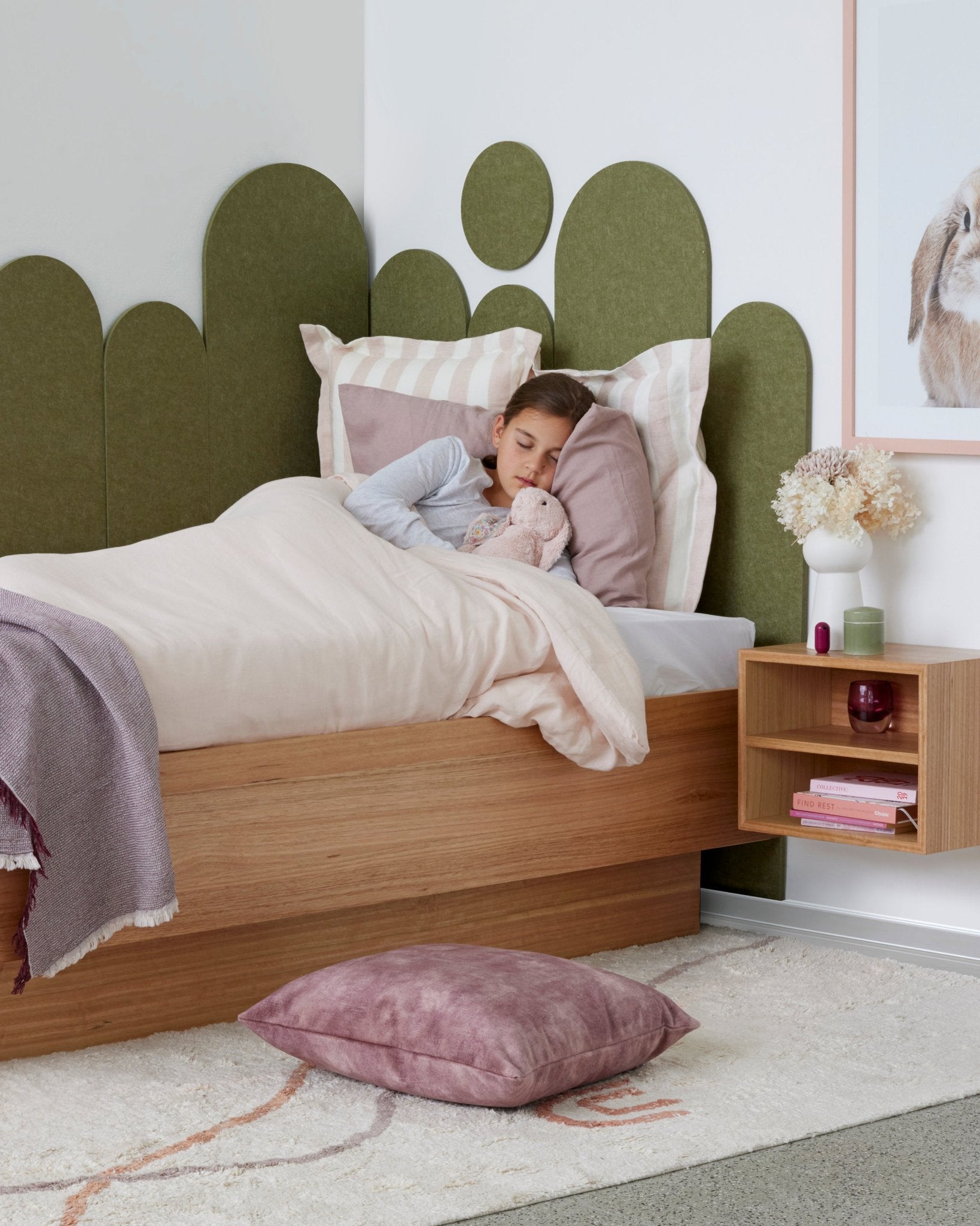A child is sleeping in a styled bedroom with a wooden base and wooden bedside table. The wall features an olive coloured felt headboard which frames the bed. The bedding to match is a soft pink with large pink striped pillowcases.