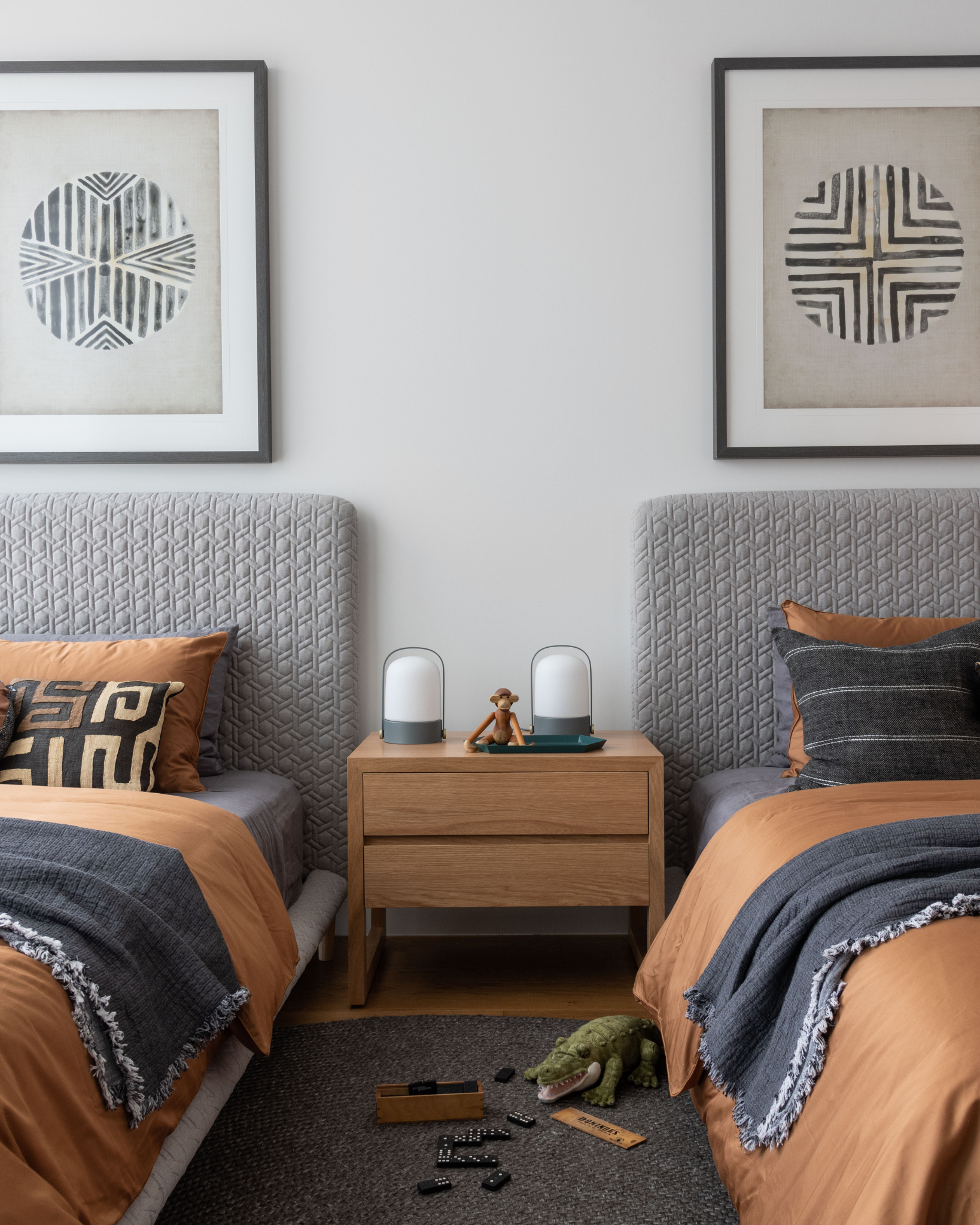 Twin bed room featuring a styled bedroom for teen boys with grey upholstered bedheads. In the middle of the beds is a wooden bedside table and the bedding is tan and black. Two geometric framed artworks are featured above both bedheads.
