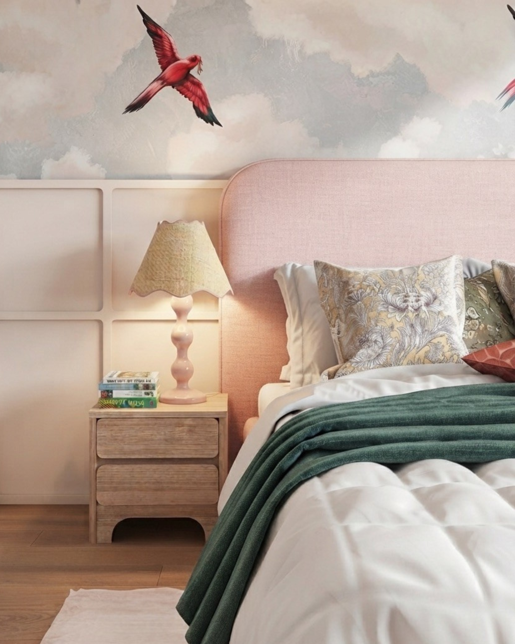 A styled bedroom scene featuring a bed with a light pink upholstered headboard, white bedding, a dark green throw blanket, a wooden nightstand with a lit table lamp, and a wall mural with a red bird and clouds.