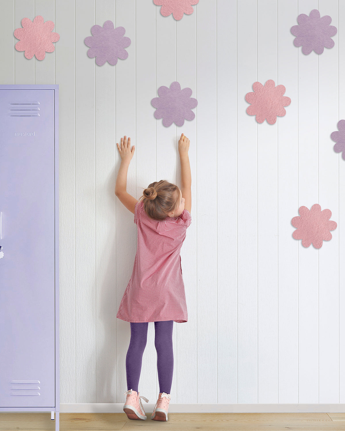 Child in a pink dress reaching up to colorful flower pinboards on a white wall next to a purple locker.