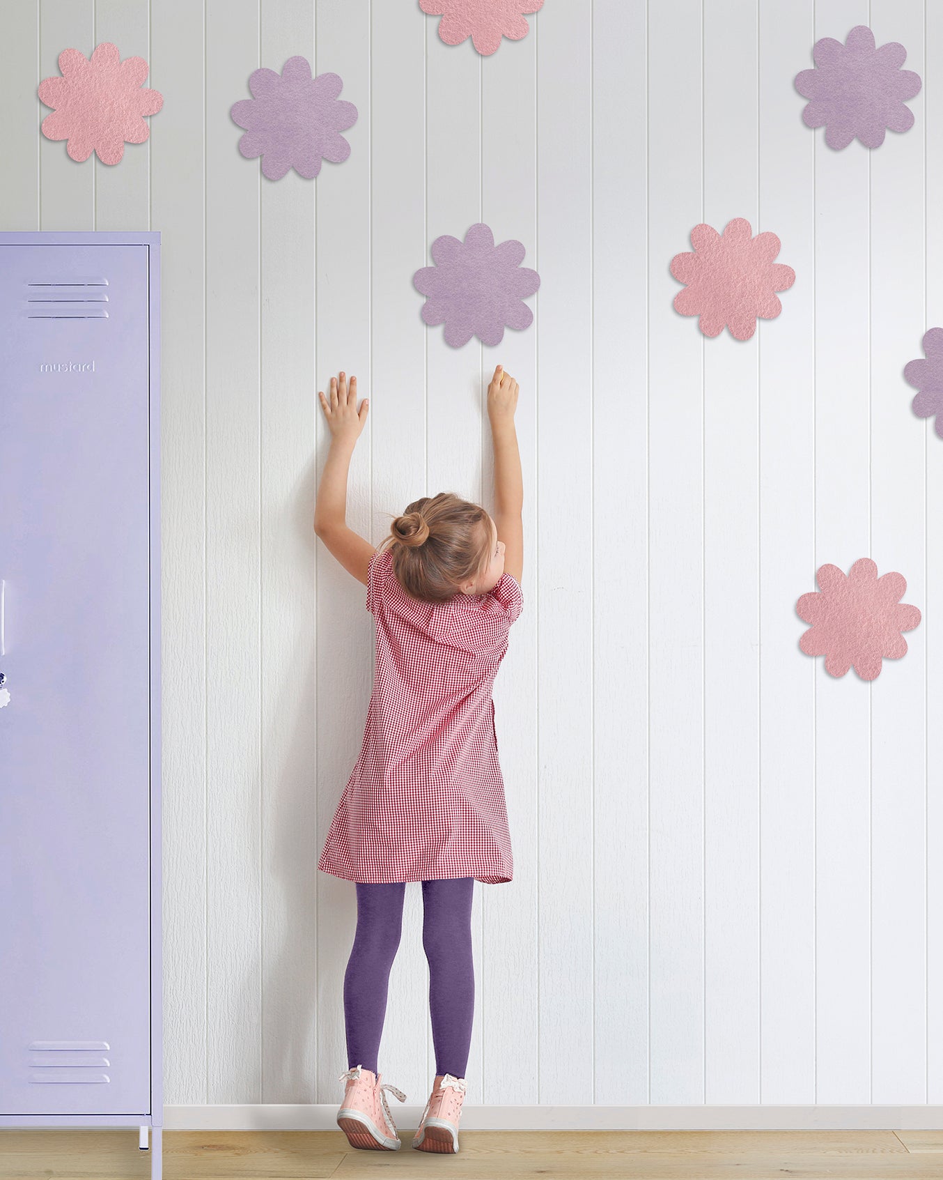 Child in a pink dress reaching up to colorful flower pinboards on a white wall next to a purple locker.
