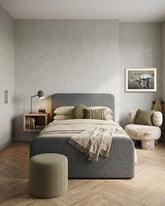 A modern bedroom featuring a grey upholstered bed with olive green bedding, a matching grey cylindrical ottoman, a floating wooden bedside table with a black desk lamp, and a fluffy armchair with a green pillow, all set against a light grey plaster wall and light wood herringbone flooring.