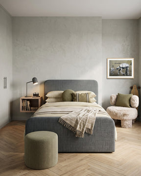 A modern bedroom featuring a grey upholstered bed with olive green bedding, a matching grey cylindrical ottoman, a floating wooden bedside table with a black desk lamp, and a fluffy armchair with a green pillow, all set against a light grey plaster wall and light wood herringbone flooring.