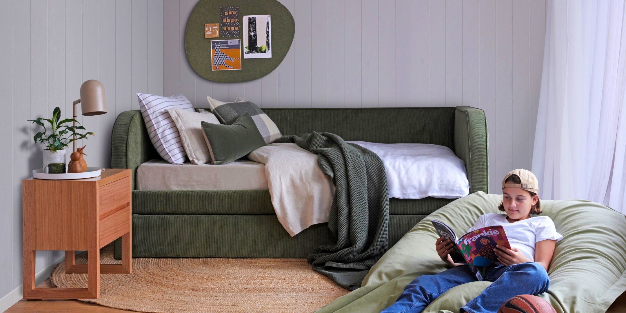 Upholstered olive green bed with a green pinboard on the wall. Timber nightstand next to the bed with a camel coloured lamp and plant. A teenager is sitting on the floor in a beanbag reading a cool magazine.