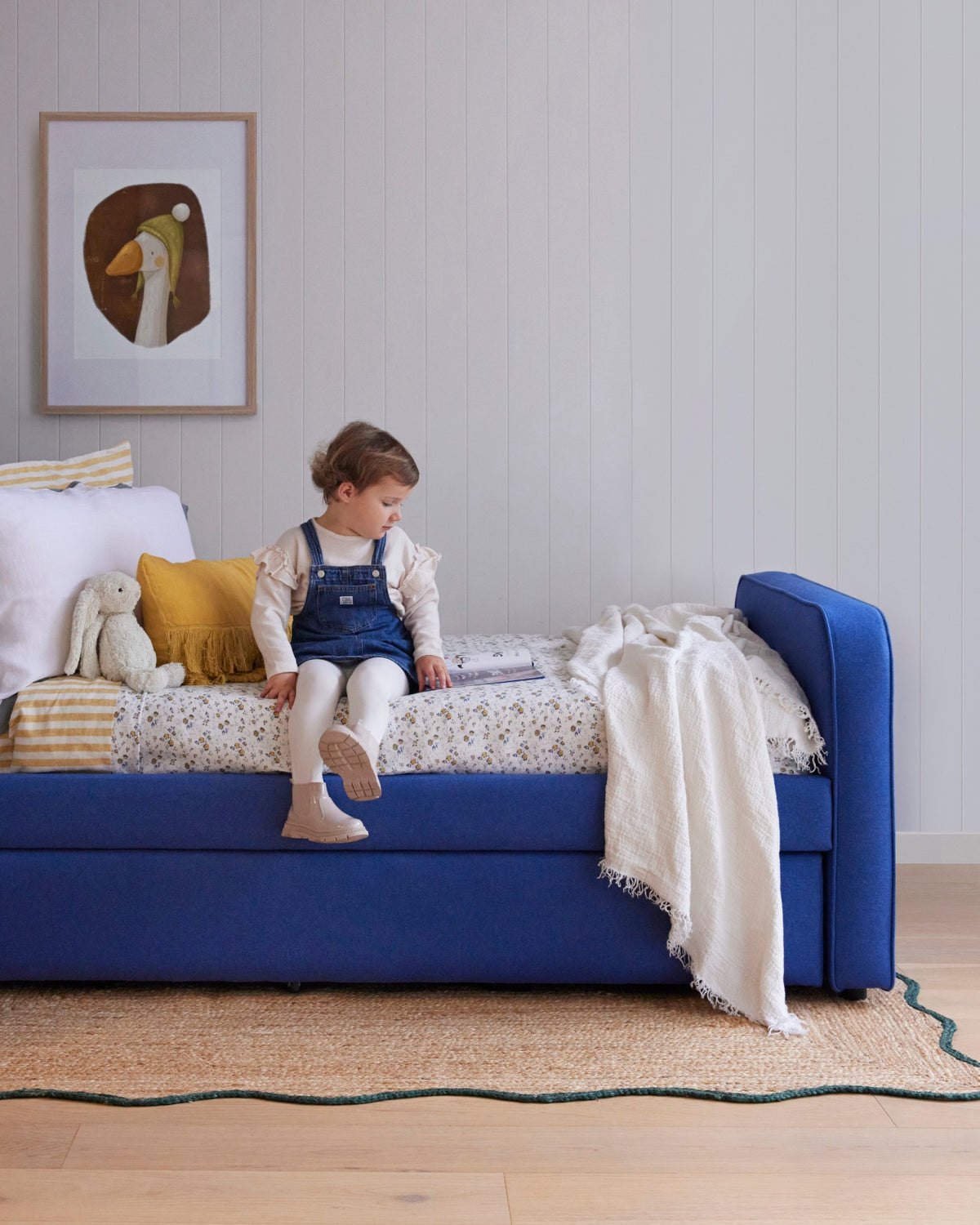 Child sitting on blue fabric bed. The Archer Upholstered Trundle is designed to sit under the Archer Upholstered Bed and rolled out when guests arrive. This trundle is perfect for children's sleepovers and guests.