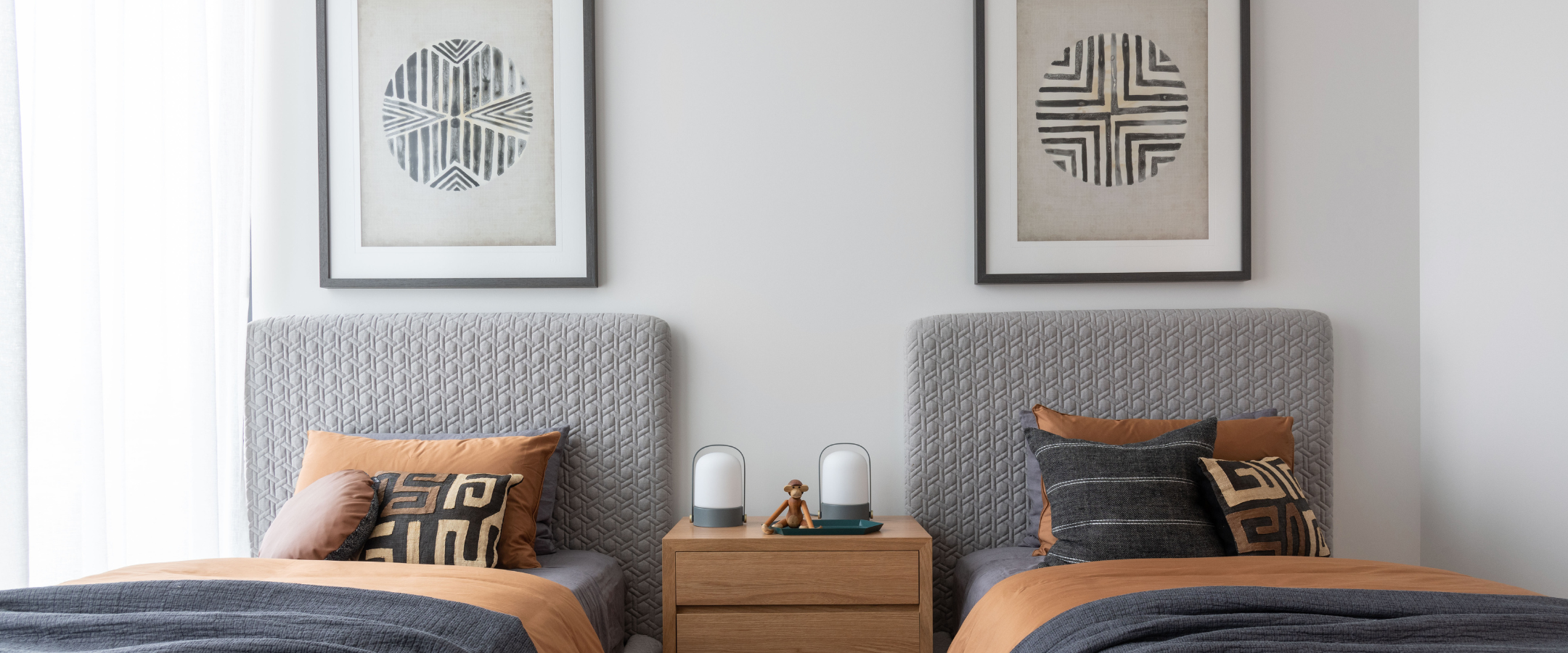 A teenager's bedroom scene showing two beds with textured gray upholstered bedheads, tan and navy bedding, patterned pillows, a small wooden bedside table, and two framed geometric wall prints above.