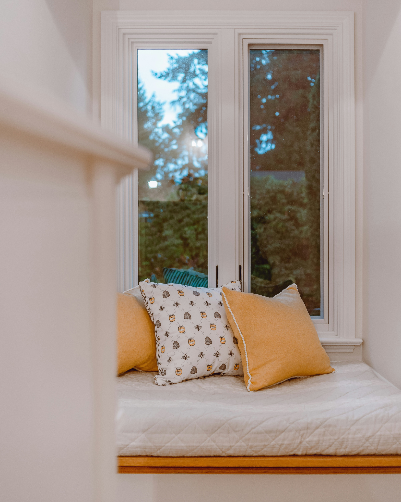 A cosy window seat with a cream quilted bench seat, styled with two mustard yellow pillows and a patterned accent pillow, set in front of a white double-hung window.