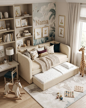 An ecru upholstered daybed with an extended trundle, styled in a bright children's bedroom with patterned pillows, a knit throw blanket, a rug, shelving, toys, and a giraffe figurine near a window.