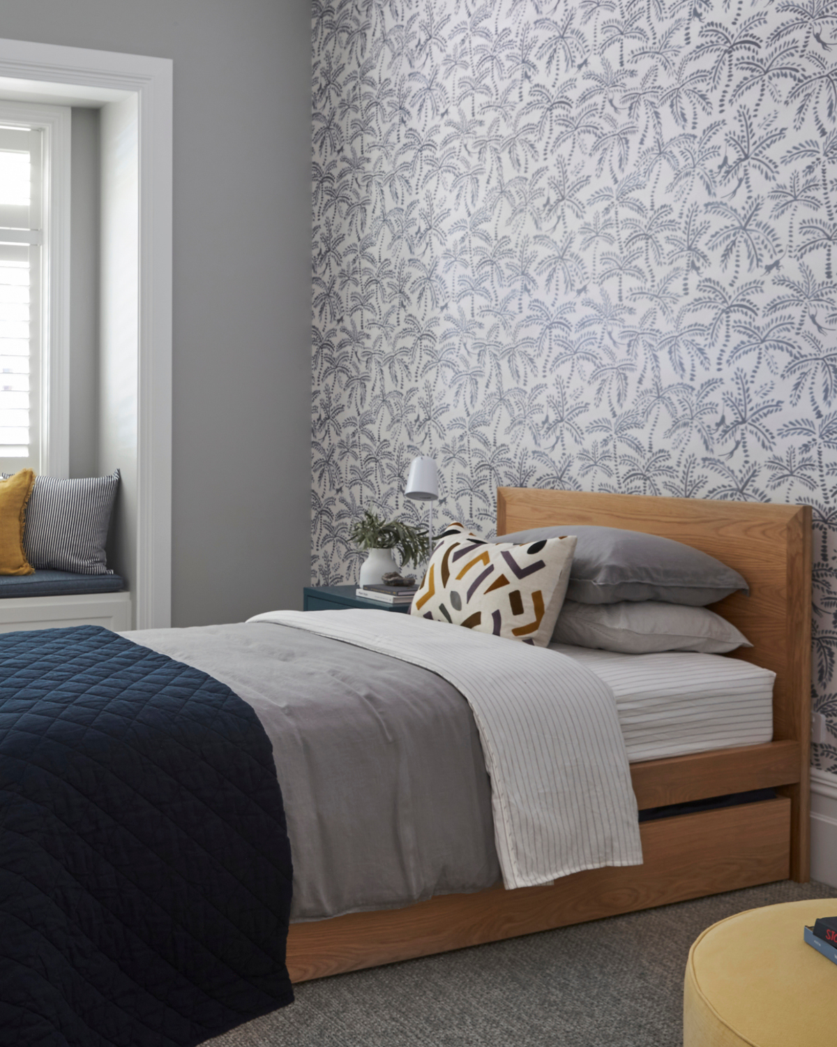 A styled bedroom interior featuring a wooden bed with trundle underneath for sleepovers. The kid's bed is set against a wall with botanical patterned wallpaper. A window with white shutters and a window seat with yellow and grey pillows are visible on the left.