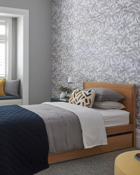 A styled bedroom interior featuring a wooden bed with trundle underneath for sleepovers. The kid's bed is set against a wall with botanical patterned wallpaper. A window with white shutters and a window seat with yellow and grey pillows are visible on the left.