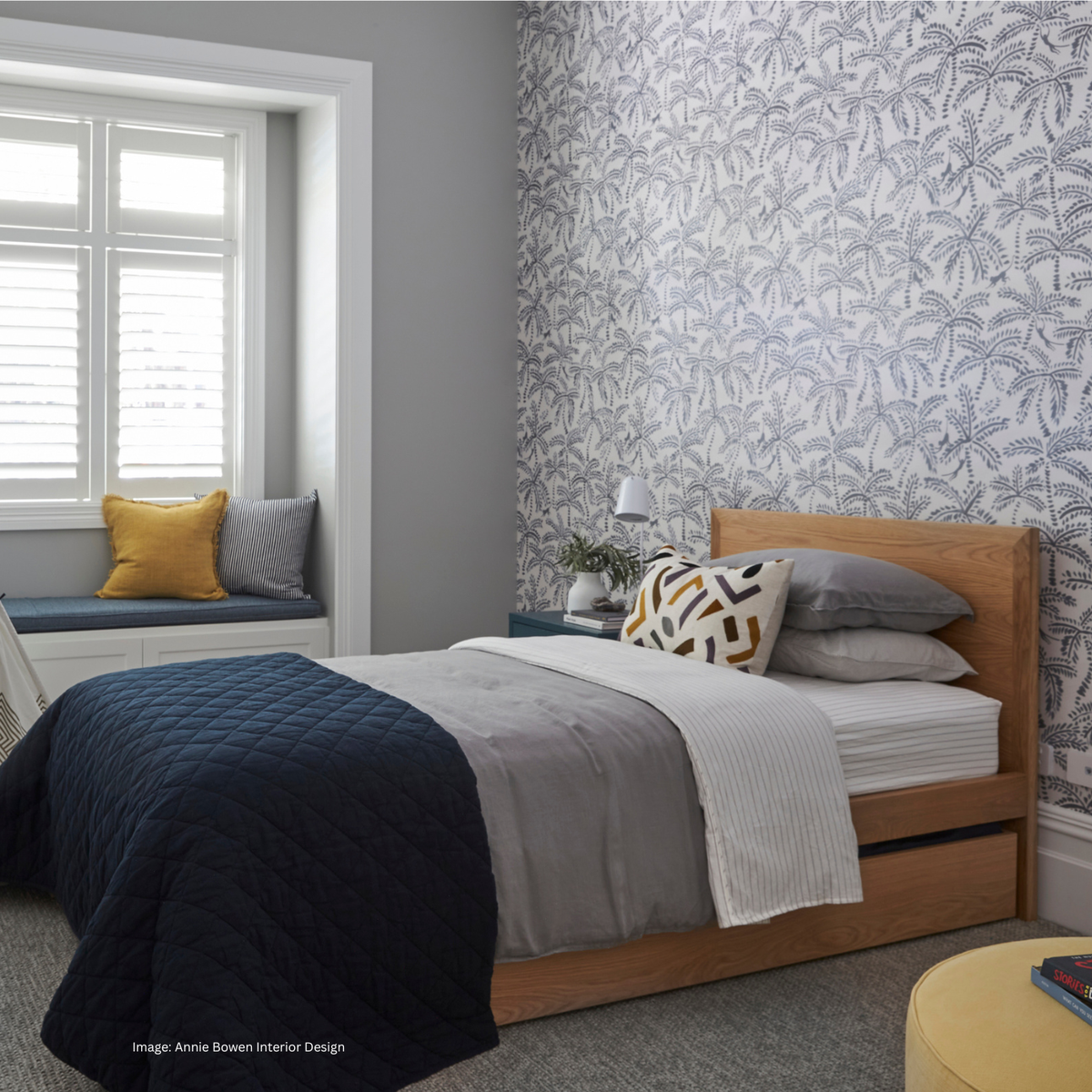A styled bedroom interior featuring a wooden bed with trundle underneath for sleepovers. The kid's bed is set against a wall with botanical patterned wallpaper. A window with white shutters and a window seat with yellow and grey pillows are visible on the left.