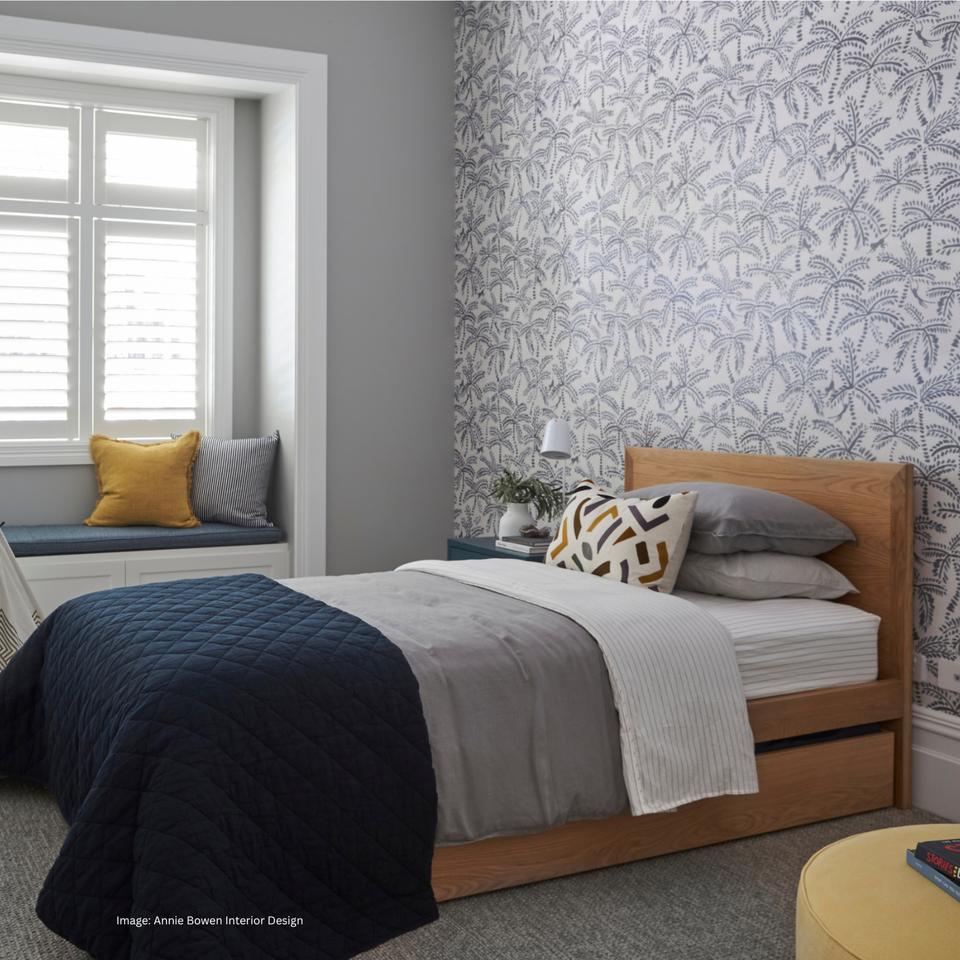 A styled bedroom interior featuring a wooden bed with trundle underneath for sleepovers. The kid's bed is set against a wall with botanical patterned wallpaper. A window with white shutters and a window seat with yellow and grey pillows are visible on the left.